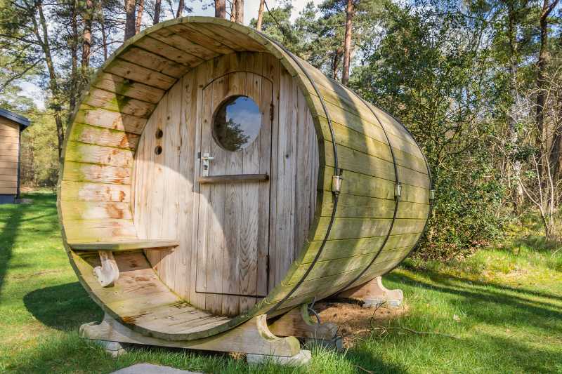 Cylindrical wooden barrel sauna with arched door and circular window, nestled among pine trees in a forest setting.