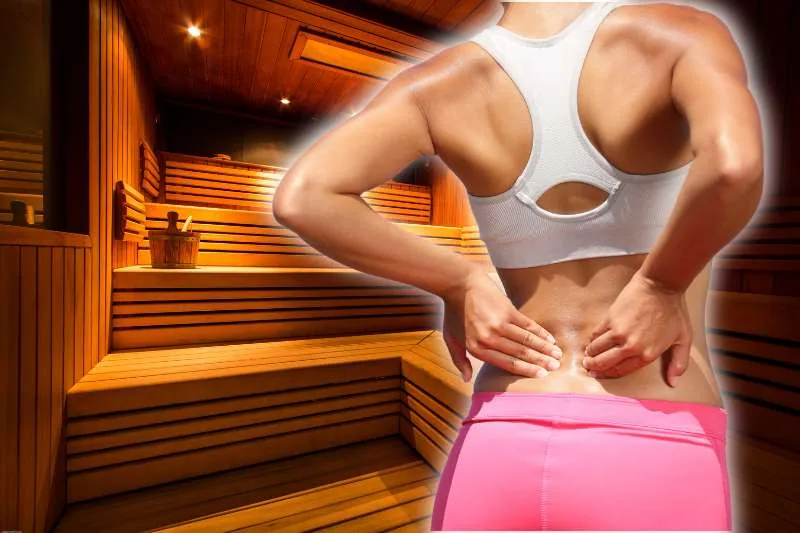 A woman in a white sports bra and pink pants holding her lower back with a wooden sauna in the background, highlighting the benefits of using a sauna for back pain relief.