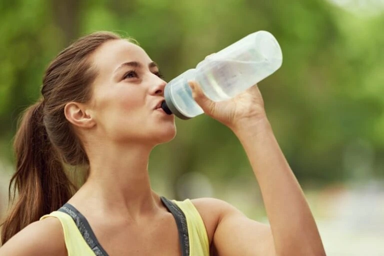 Woman in a yellow tank top drinking water from a bottle, emphasizing hydration for sauna use.