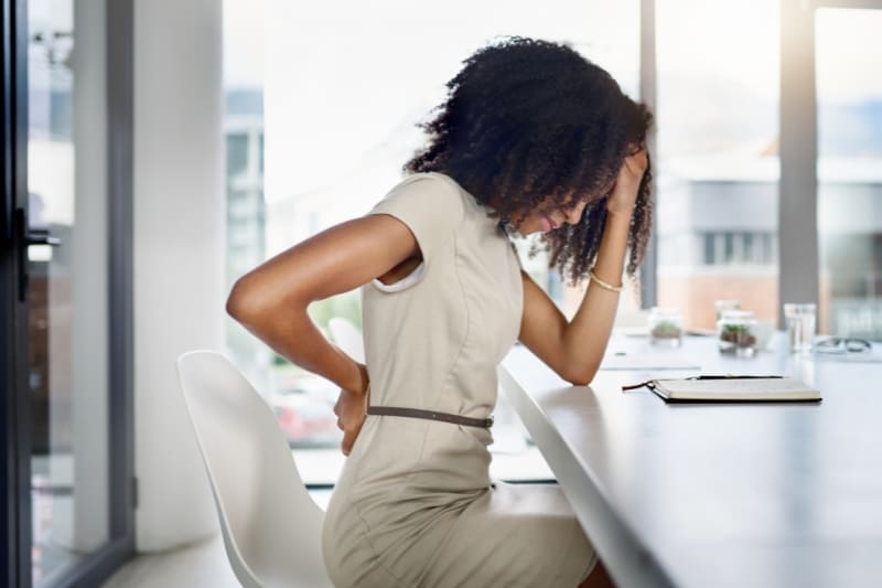 A woman sitting at a desk in an office, holding her lower back in pain while leaning forward.