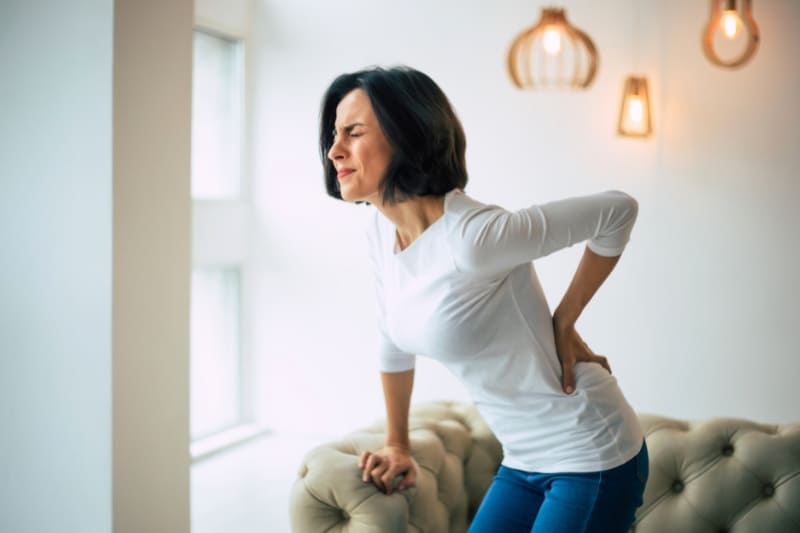 A woman in a white shirt and blue pants sitting on a couch, holding her lower back in pain.