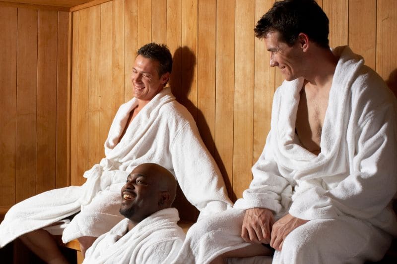 Three men of different ethnicities smile and relax in a sauna wearing white bathrobes, enjoying the heat and camaraderie.