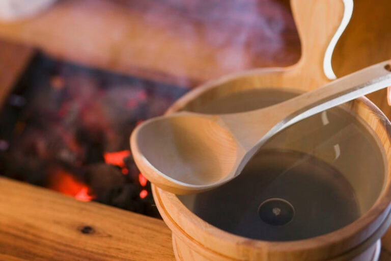 A wooden ladle rests on a bucket of water in a sauna, ready to pour water onto hot sauna rocks and generate steam, increasing the humidity and perceived heat.