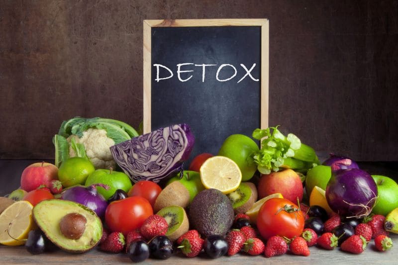 A variety of colorful fruits and vegetables, including apples, berries, and avocados, are displayed in front of a chalkboard with the word "DETOX" written on it, promoting the concept of a healthy detox diet.