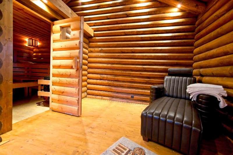 A home sauna with a wooden door set in a log cabin-style room, featuring a lounge chair and towels for relaxation.