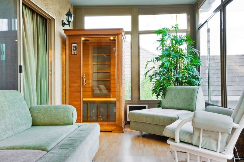 An infrared sauna placed in a bright sunroom with comfortable seating and potted plants.