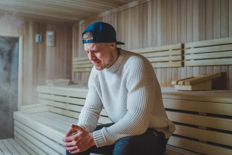 Man sitting in a sauna wearing a sweater and cap, visibly uncomfortable, wondering why is my sauna not heating up