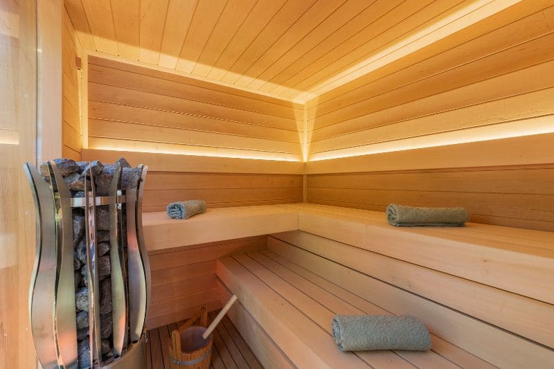 Modern sauna featuring warm wood paneling, LED accent lighting, tiered benches, and a stylish heater filled with stones