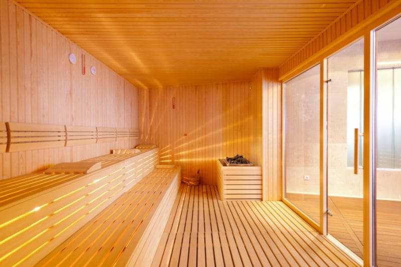 Modern sauna with light wood paneling, benches, a heater, and a glass door.