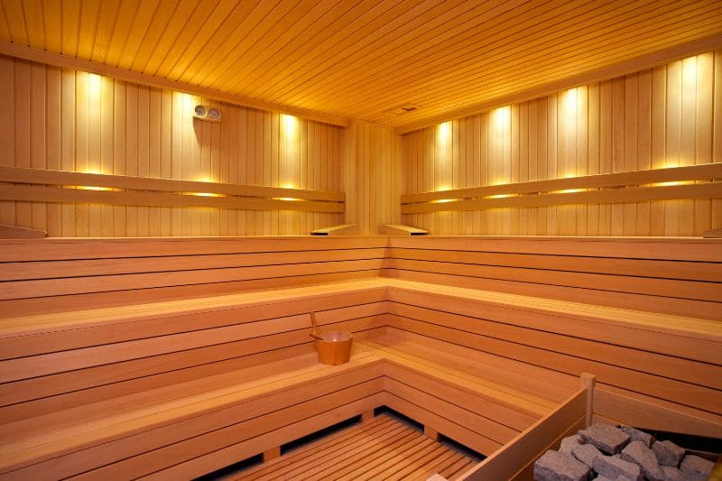 A modern sauna interior with a built-in electric heater and a wooden bucket on the floor. The sauna features a spacious seating area and a well-lit interior.