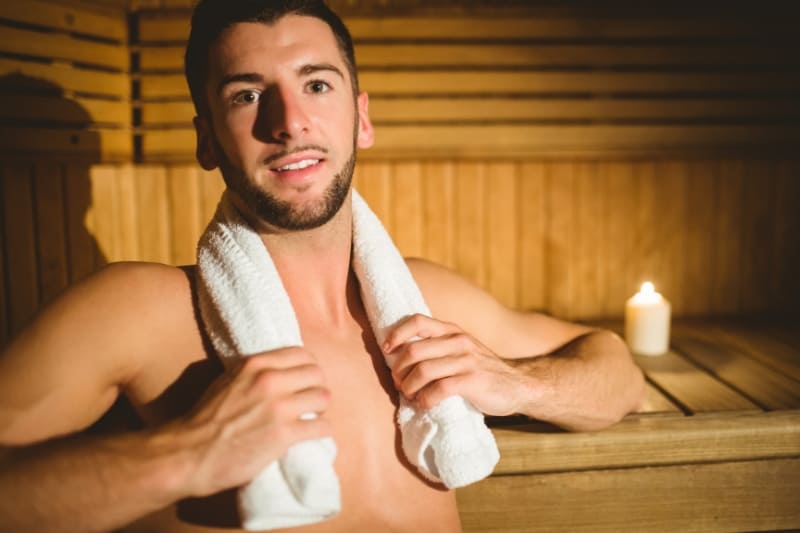 A man relaxing in a sauna before a workout, holding a towel around his neck. The warm wooden interior and soft lighting create a calming atmosphere.