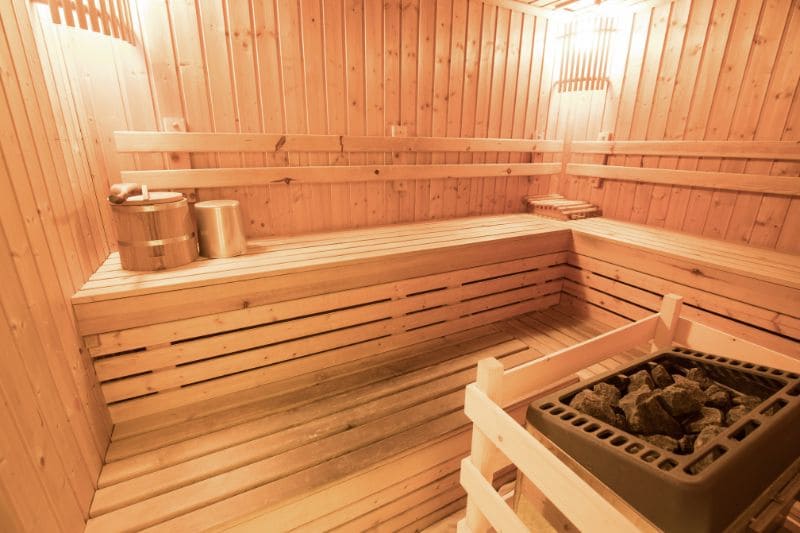 A sauna interior with wooden benches, a traditional rock heater, a bucket, and a ladle.