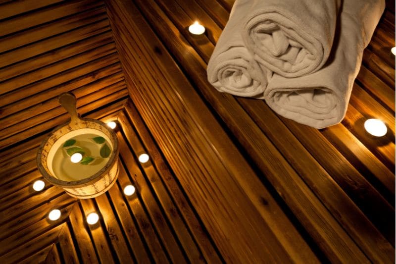 A sauna with candles lit around the floor and two white towels rolled up in the corner. The wooden walls create a warm and relaxing atmosphere.