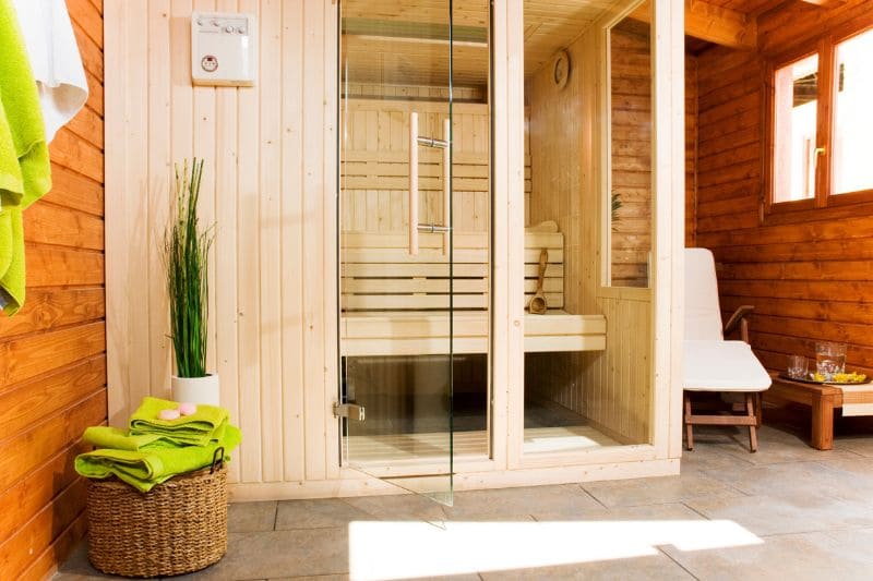 A traditional home sauna with wooden walls, a glass door, and benches inside. Towels and a plant add a decorative touch.