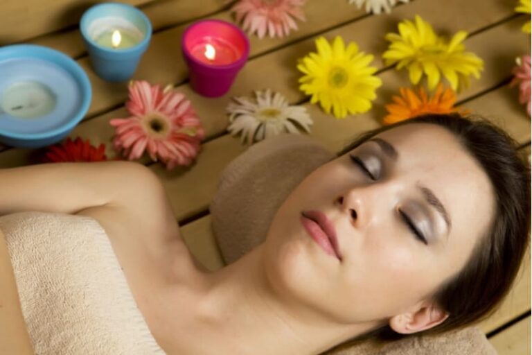 A woman with her eyes closed enjoys the relaxing benefits of aromatherapy in sauna, surrounded by candles and colorful flowers.