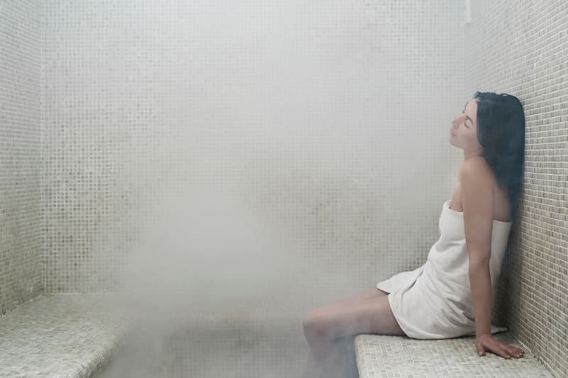 Young woman in a towel relaxing in a steam room filled with dense steam, highlighting the soothing benefits of using a steam room for cold relief.