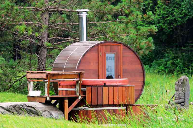 Barrel Sauna in a natural outdoor forest setting, featuring cedar wood construction, glass door, and small porch with seating.