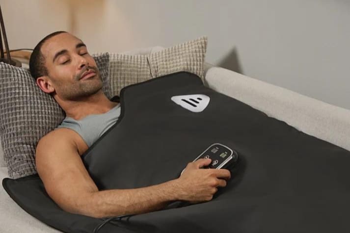 A man relaxing on a couch, covered by a black Lifepro sauna blanket, holding a remote control.