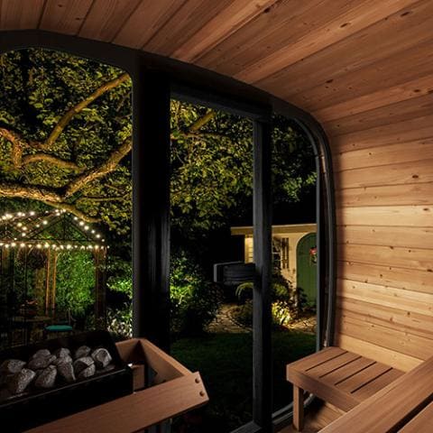 Nighttime view from inside the SaunaLife 4-Person Cube-Series Outdoor Home Sauna Kit CL5G, featuring a large glass panel overlooking an illuminated garden, demonstrating the sauna's panoramic outdoor views and wooden interior.