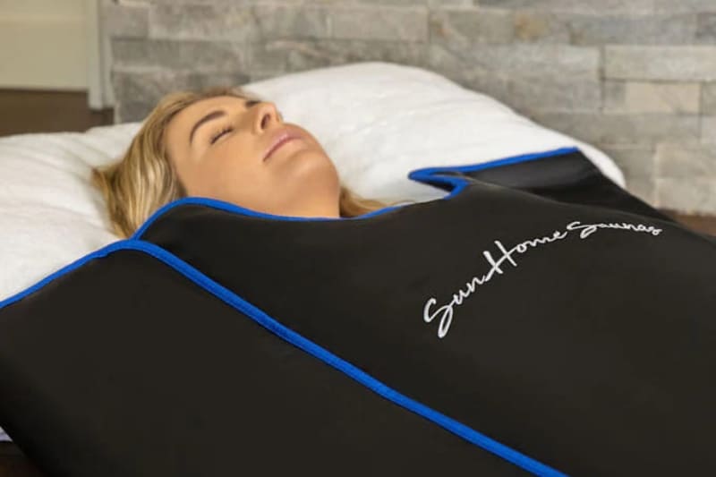 Close-up of a blonde woman's face as she rests inside a black Sun Home sauna blanket with blue edges and white logo.