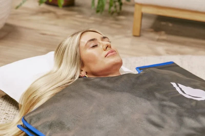 A blonde woman lying relaxed on the floor with her eyes closed, enveloped in a sleek black Sun Home infrared wrap experiencing sauna blanket benefits.