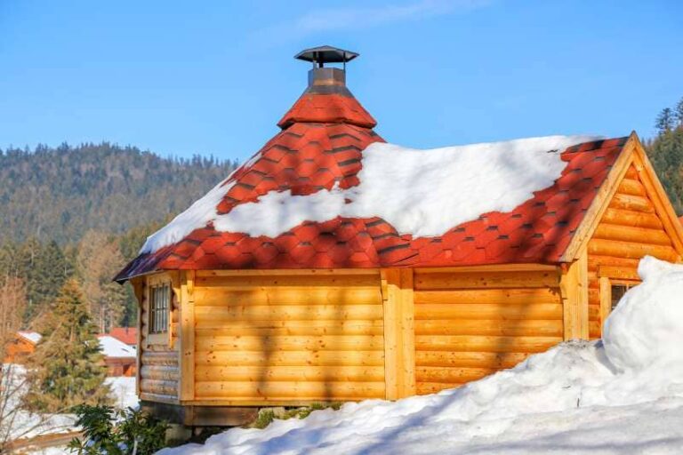 Traditional Outdoor wooden Sauna with bright orange shingle roof partially covered in snow, set against a mountainous winter landscape.