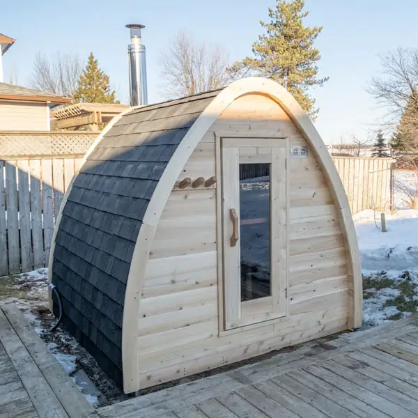 Compact wooden barrel sauna with glass door and black shingled roof, placed on a deck in a snowy backyard setting with visible chimney.