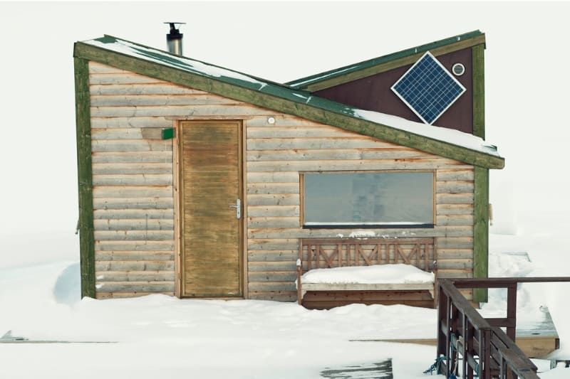 Rustic wooden outdoor sauna cabin with slanted roof, solar panel, and small porch, surrounded by snow, showcasing sustainable design for winter relaxation.