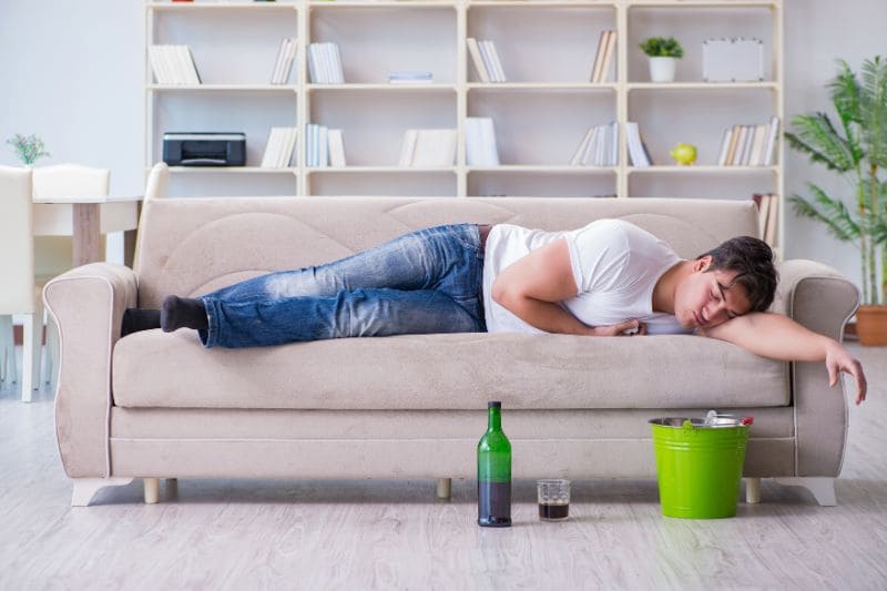 Hungover man sleeping on couch with empty bottle and bucket nearby