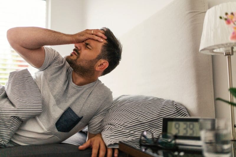 Man experiencing hangover symptoms, holding head in pain while sitting in bed