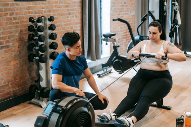 Personal trainer guiding overweight woman using rowing machine in gym, focusing on fitness and exercise