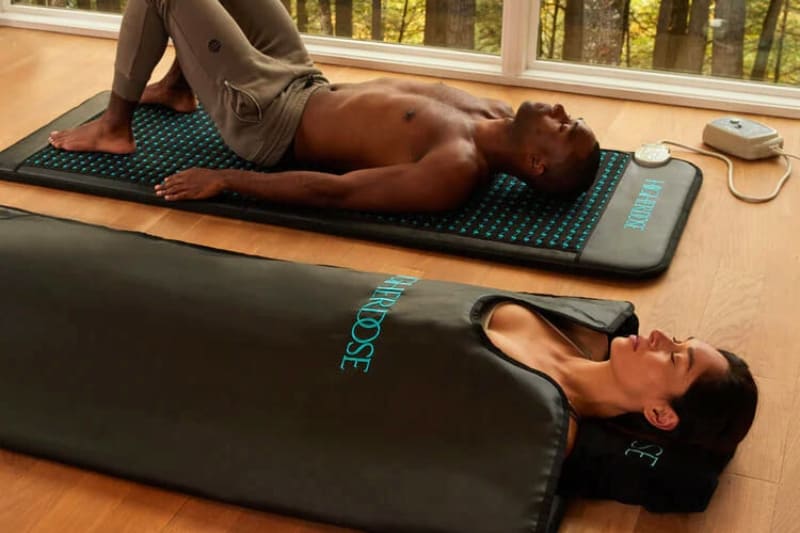 Two people using HigherDose wellness products. One person lies on a blue-lit mat, while another is inside a black sauna blanket labeled "HigherDose".