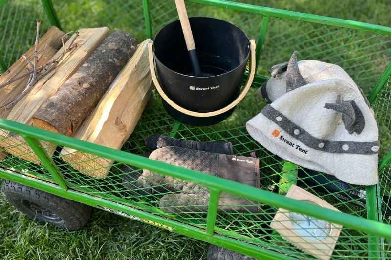 Sweat Tent Sauna accessories including firewood, bucket, ladle, and sauna hat on garden cart