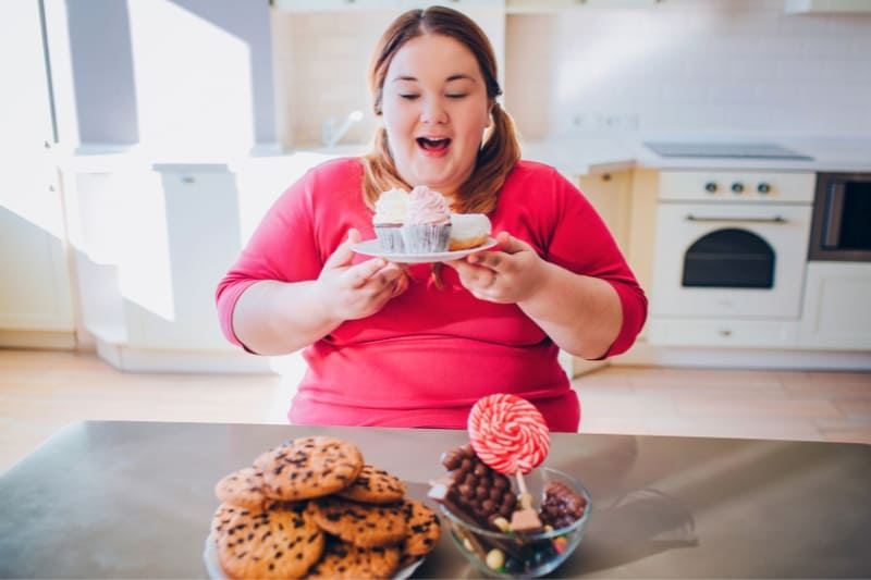 Overweight woman excitedly eating cupcakes with various sweets on table, illustrating poor dietary choices