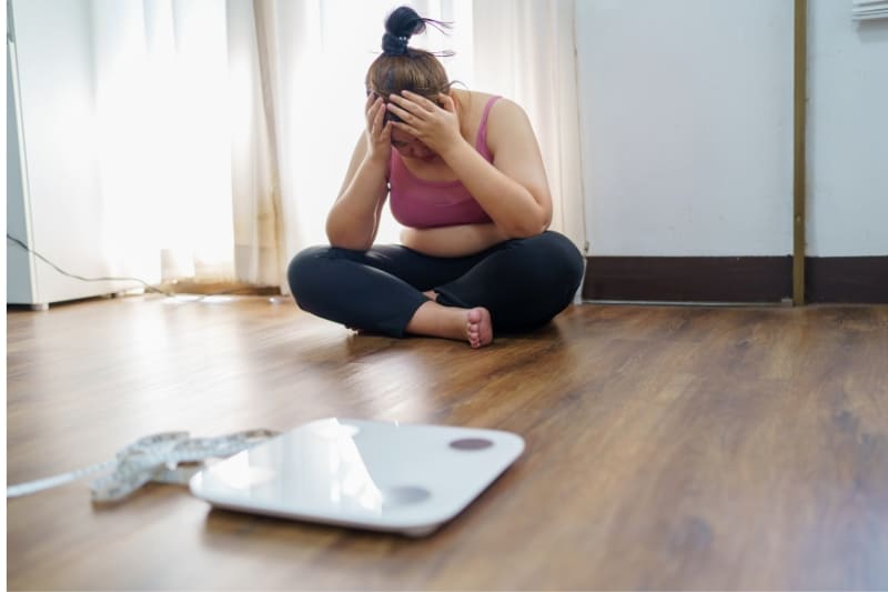 Frustrated woman sitting on floor with scale nearby, depicting emotional challenges of weight management