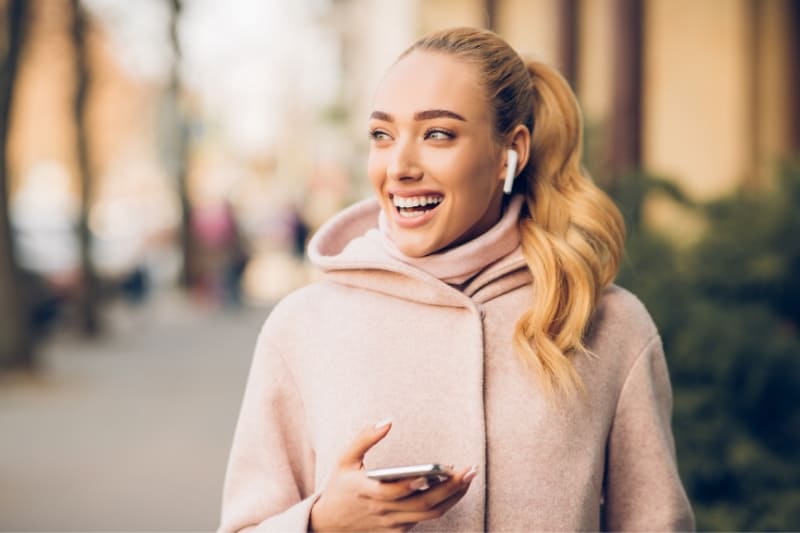 Smiling blonde woman wearing AirPods and holding smartphone while walking outdoors