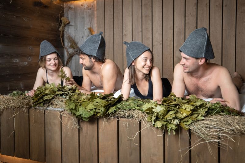 Four people wearing sauna hats relaxing in a rustic sauna with leafy branches