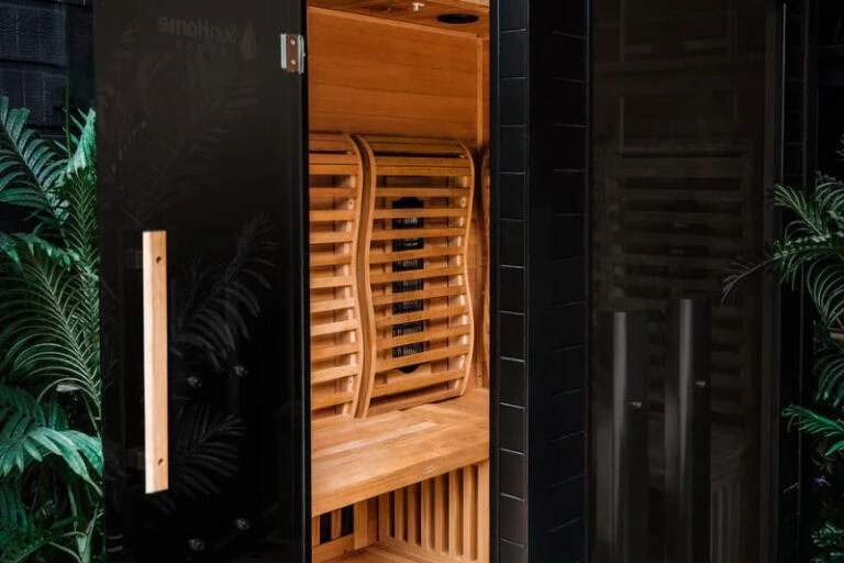 Exterior view of an infrared sauna with open door, showing wooden interior and surrounded by plants