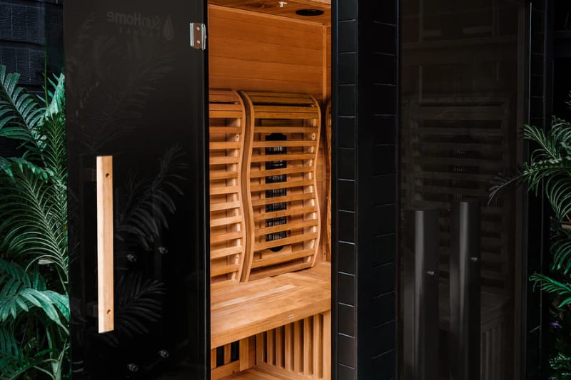 Exterior view of an infrared sauna with open door, showing wooden interior and surrounded by plants