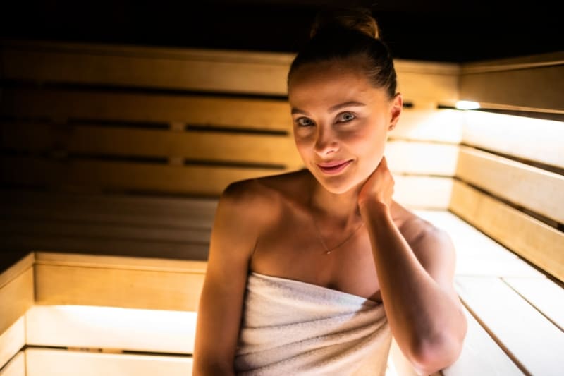 Woman wrapped in white towel enjoying warm atmosphere of illuminated sauna