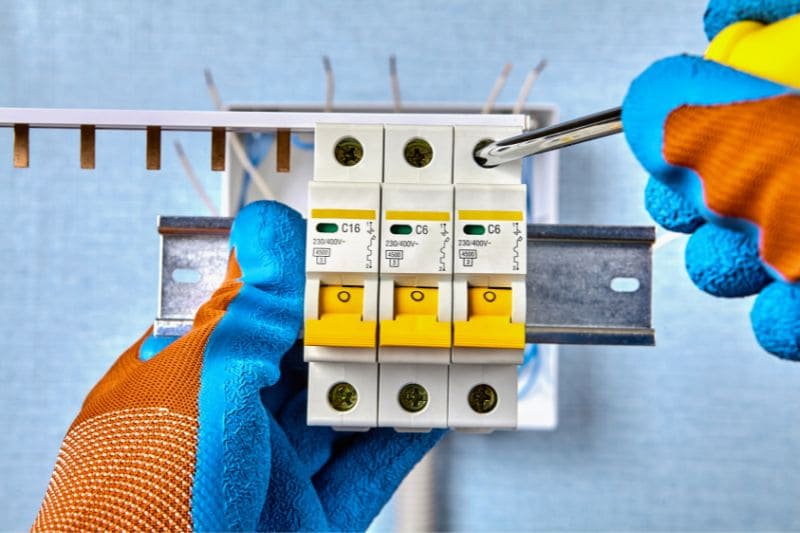 Licensed electrician in protective gloves installing circuit breakers on DIN rail, demonstrating professional electrical work required for safe sauna installation