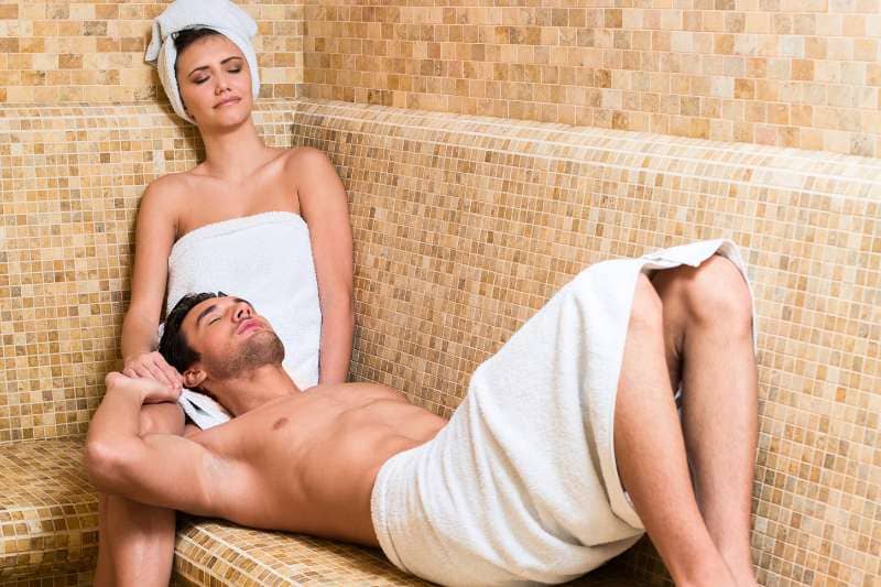 Couple relaxing in luxury spa steam room with tiled beige mosaic walls, wrapped in white towels, resting on heated bench during wellness treatment