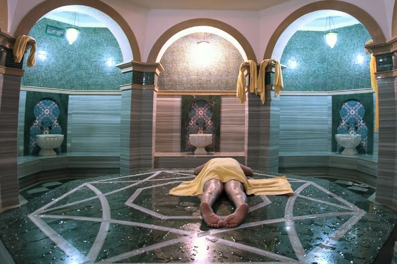 Traditional Turkish hammam interior with turquoise mosaic tiles, arched alcoves, marble massage platform, steam room with decorative geometric floor pattern and person lying covered in yellow towel