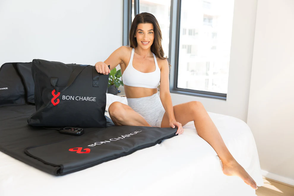 Woman in athletic wear demonstrating BON CHARGE Infrared Sauna Blanket setup on bed in modern bedroom