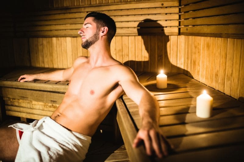 Young man relaxing in traditional wooden sauna with candles, exposing body to therapeutic heat