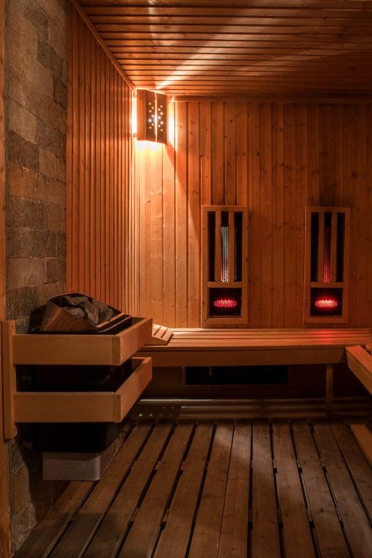 Interior of a hybrid sauna featuring both infrared heating panels and electric sauna heater, with stone wall accent and tiered wooden benches illuminated by warm lighting