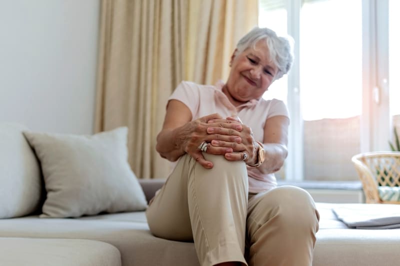 Senior woman experiencing knee pain from osteoarthritis while sitting on couch, holding her knee in discomfort