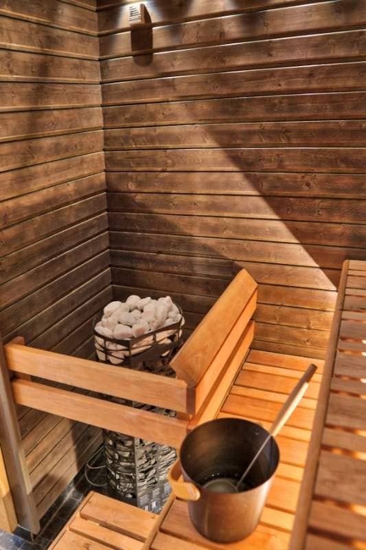 Traditional steam sauna interior with dark wood paneling, wooden benches, rock heater tower, and water bucket with ladle