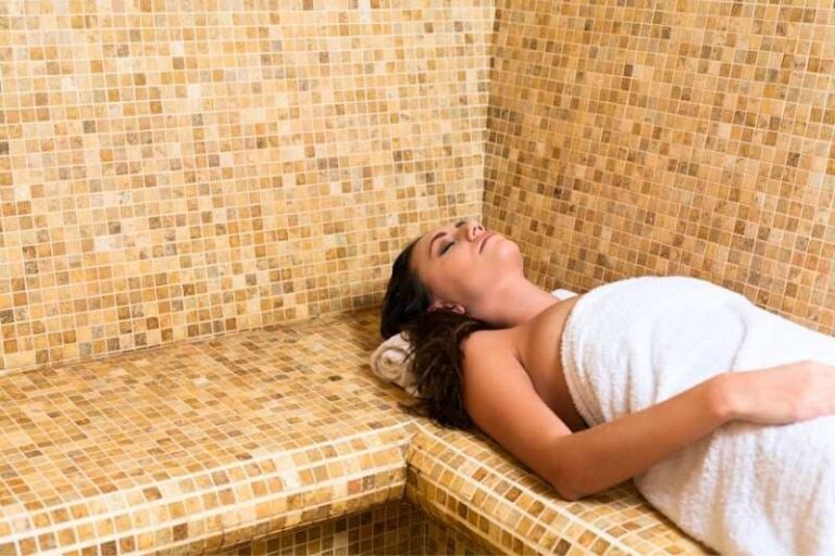 Woman relaxing on mosaic-tiled thermal bench in luxury spa steam room, wrapped in white towel, eyes closed in peaceful meditation