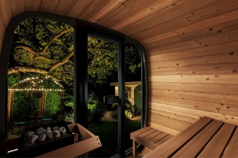 Interior view of the Saunalife Cube Series outdoor home sauna featuring a modern wood-paneled design, a cozy bench seating area, and a glass door opening to a beautifully lit backyard with string lights and lush greenery at night.